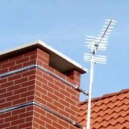 Antena telewizyjna zamontowana na kominie z czerwonej cegły na tle błękitnego nieba i fragmentu dachu z czerwonej dachówki karpiówki.