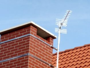 Antena telewizyjna zamontowana na kominie z czerwonej cegły na tle błękitnego nieba i fragmentu dachu z czerwonej dachówki karpiówki.