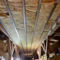 Izolacja poddasza wełną mineralną, widoczne metalowe profile mocujące, drewniana konstrukcja dachu, materiały budowlane składowane w tle.