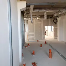 Wnętrze w trakcie prac wykończeniowych: widoczne metalowe profile ścian działowych, instalacja wentylacyjna pod sufitem, rury kanalizacyjne wystające z wylewki oraz częściowo zamontowane płyty...