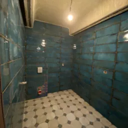 Wnętrze łazienki w trakcie remontu z ułożonymi turkusowymi płytkami na ścianach i wzorzystą podłogą, widoczne instalacje wodne i surowy betonowy sufit z wiszącą żarówką.