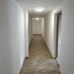 Długi korytarz z nową podłogą ułożoną w jodełkę, świeżo pomalowane białe ściany i okrągłe lampy sufitowe, widok z perspektywy.