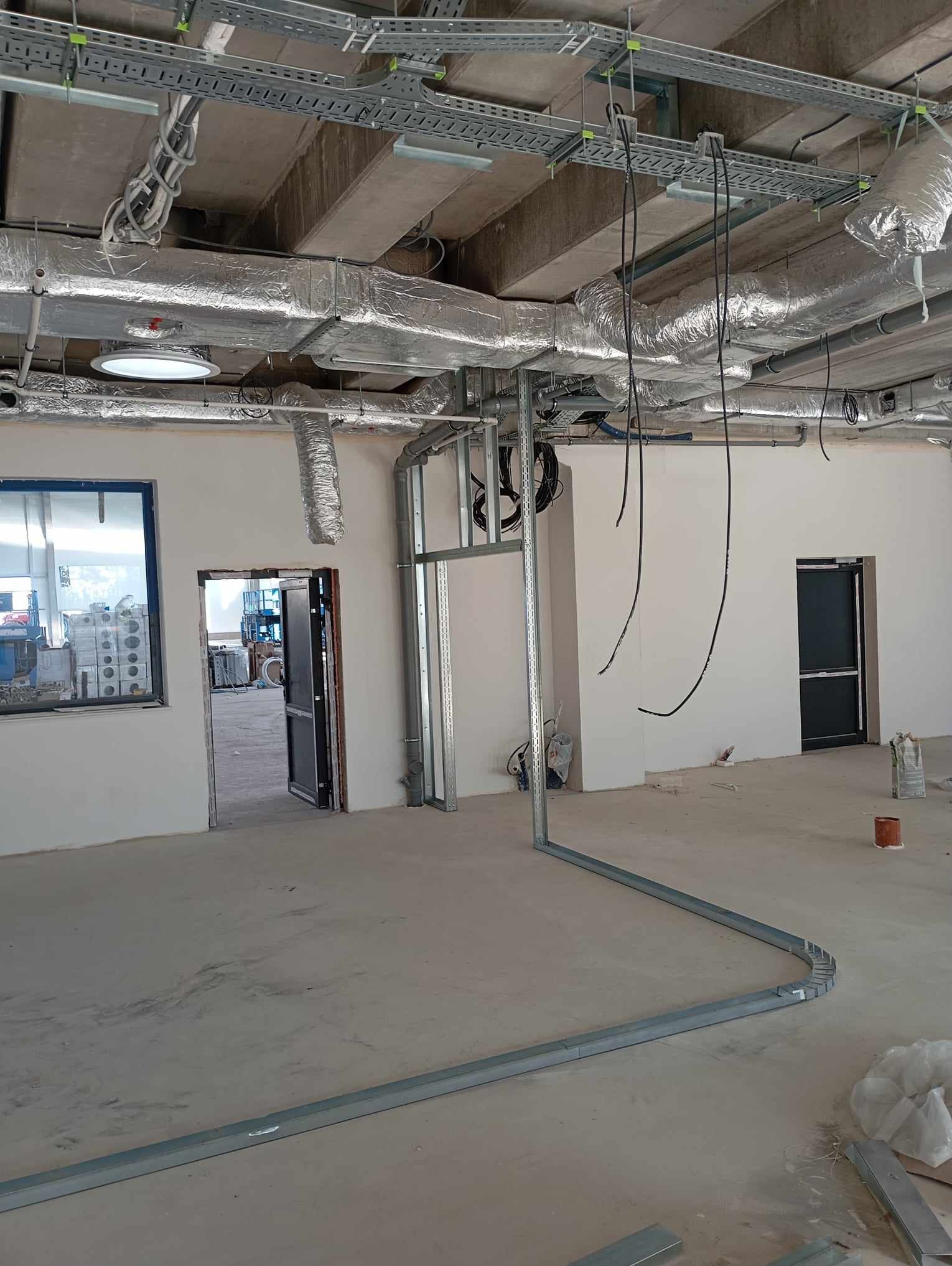 Wnętrze w trakcie prac wykończeniowych: widoczne instalacje wentylacyjne pod sufitem, metalowe profile na ścianach i podłodze, przygotowane do zabudowy, surowy beton na podłodze, otwory drzwiowe...