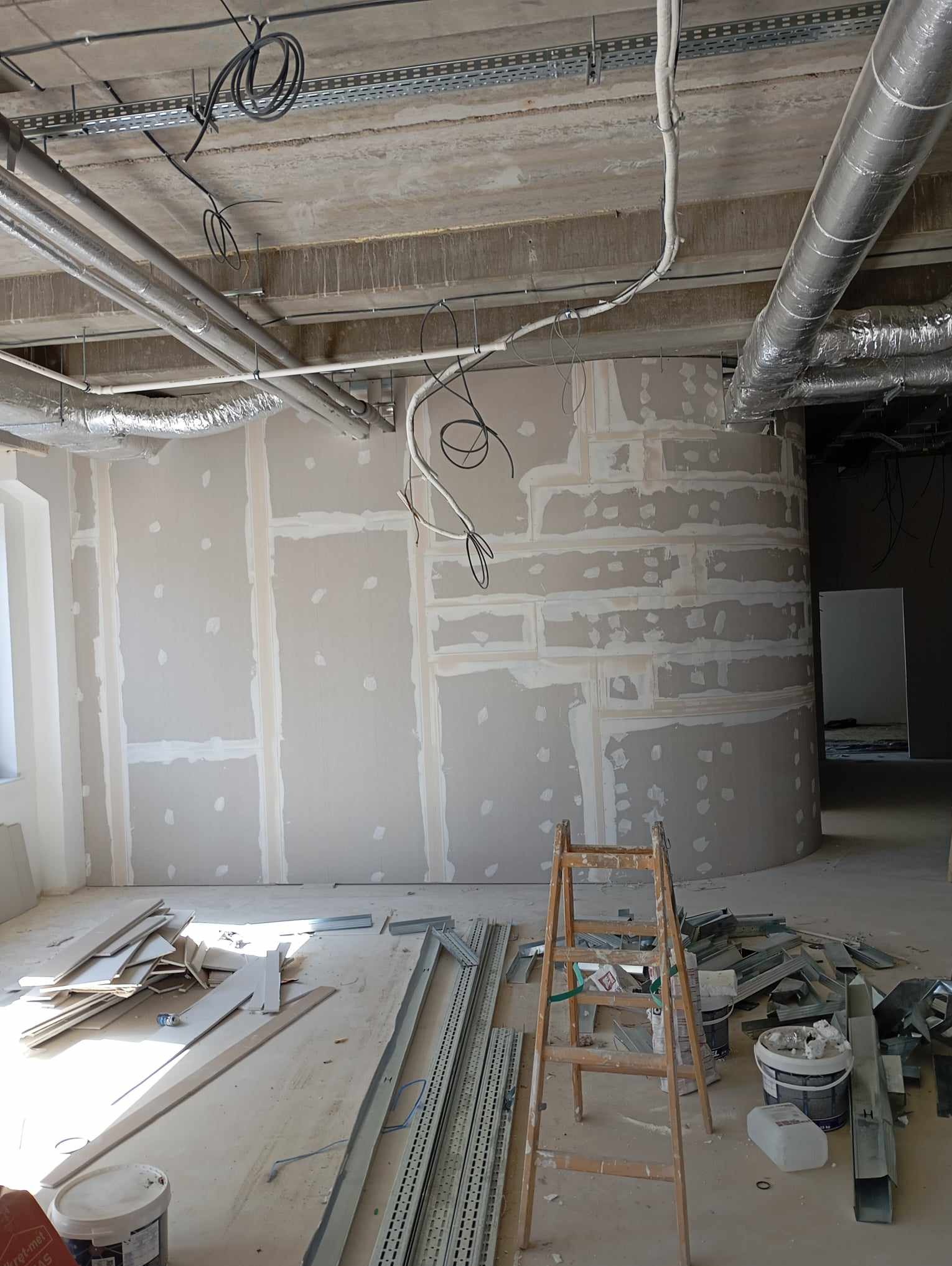 Wnętrze w trakcie prac wykończeniowych: ściany z płyt kartonowo-gipsowych z widocznymi łączeniami, metalowe profile, drabina malarska, puszki z farbą i instalacje pod sufitem.
