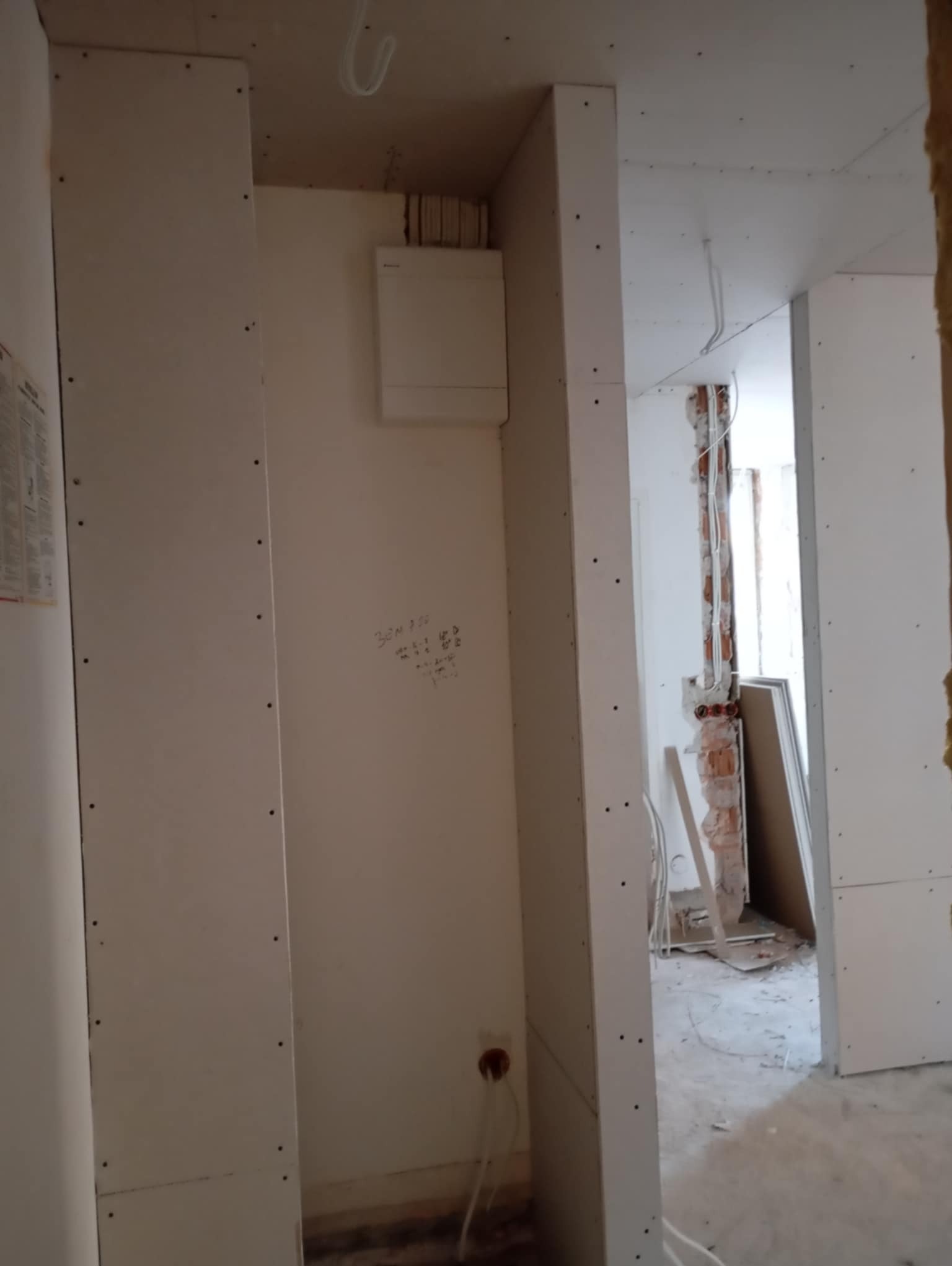 Wnętrze w trakcie prac wykończeniowych: montaż płyt na ścianach, widoczne instalacje elektryczne, fragment odsłoniętej cegły, notatki na ścianie.
