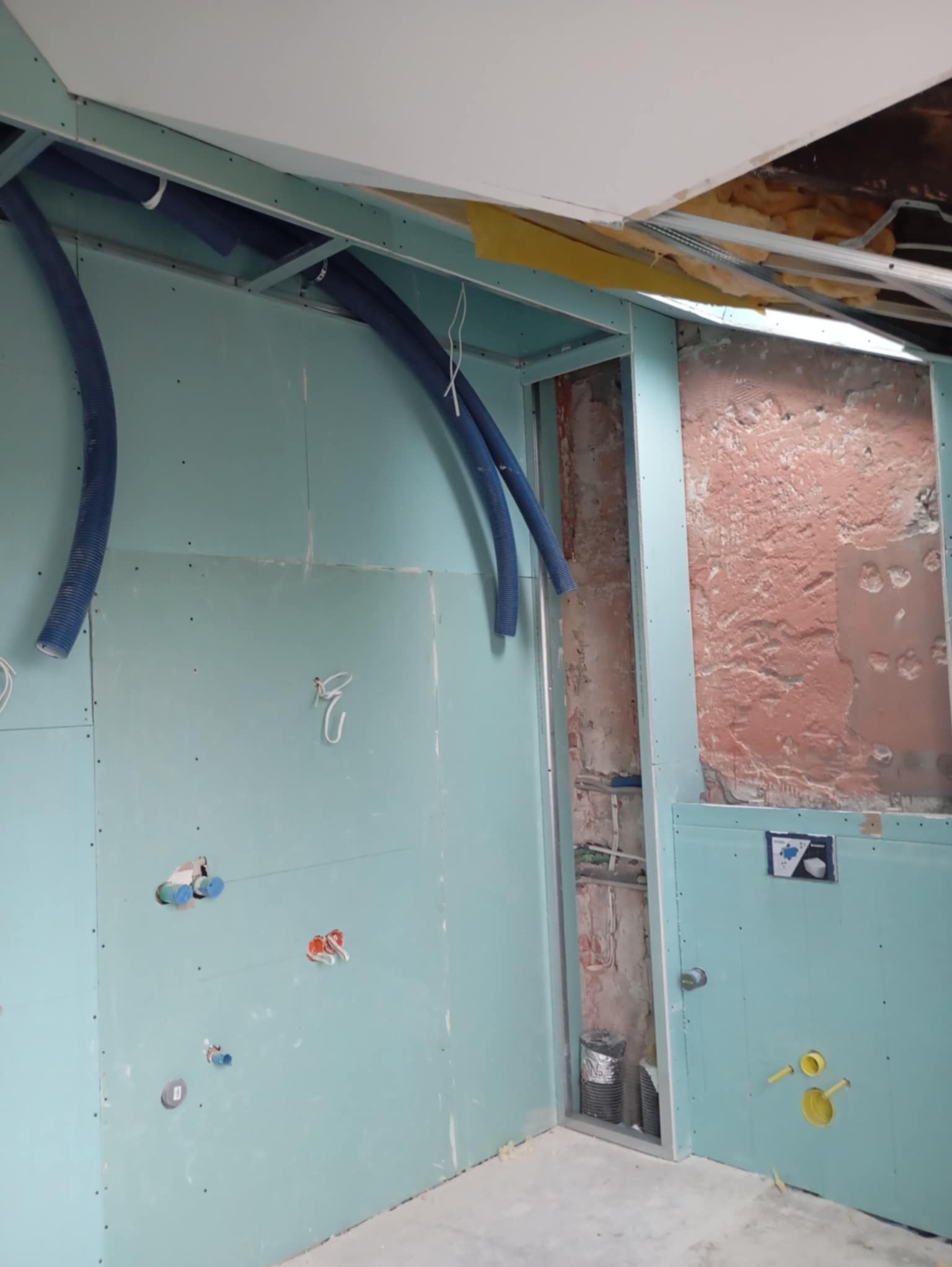 Wnętrze w trakcie prac wykończeniowych: niebieskie rury wentylacyjne prowadzone pod sufitem, ściany obłożone płytami gipsowo-kartonowymi, widoczne instalacje elektryczne i hydrauliczne w ścianach...