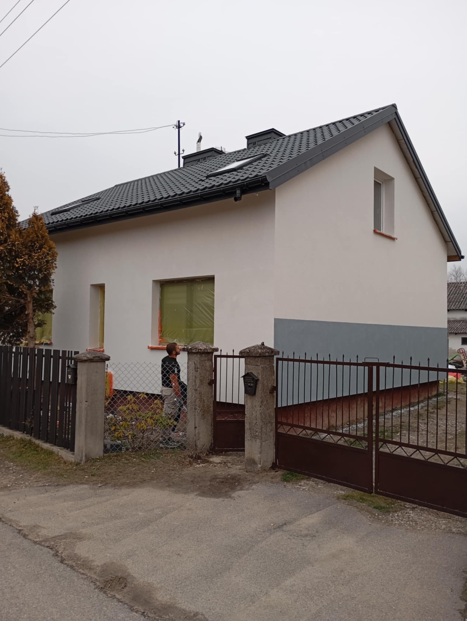 Dwukondygnacyjny dom jednorodzinny z szarą podmurówką i białymi ścianami, okna zabezpieczone folią, widoczny pracownik w trakcie pracy przy elewacji, szara dachówka, metalowe ogrodzenie.