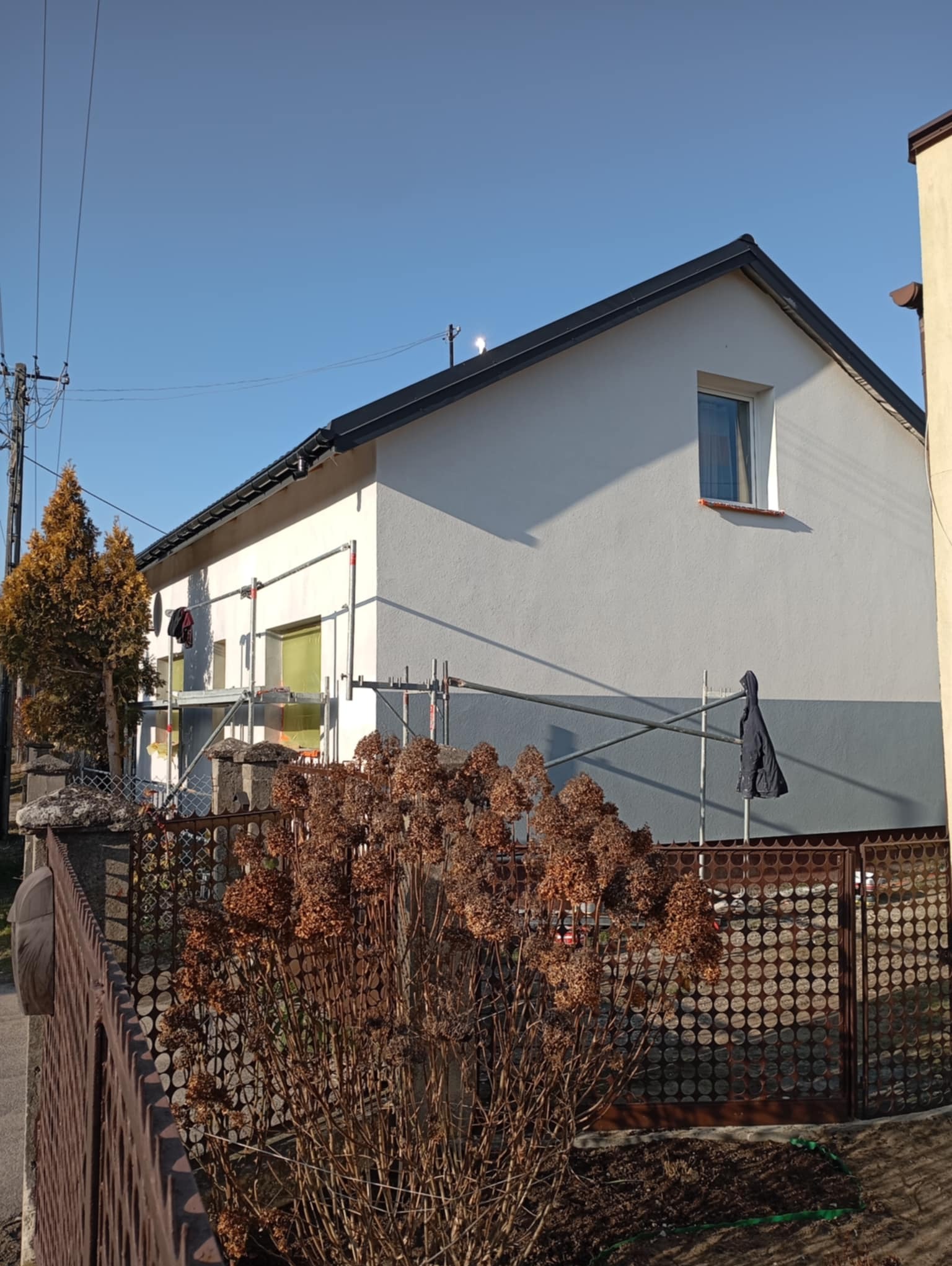 Dom w trakcie malowania elewacji, widoczne rusztowania, częściowo pomalowana ściana na biało, dolna część szara, zabezpieczone okna folią, w tle błękitne niebo.
