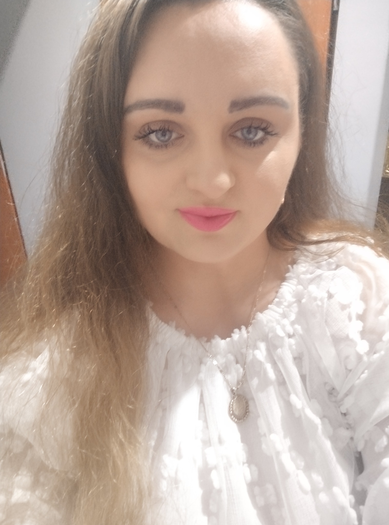 Portret kobiety z długimi, jasnymi włosami, ubranej w białą bluzkę z koronkowymi detalami i srebrny naszyjnik z owalną zawieszką. Usta pomalowane na różowo.