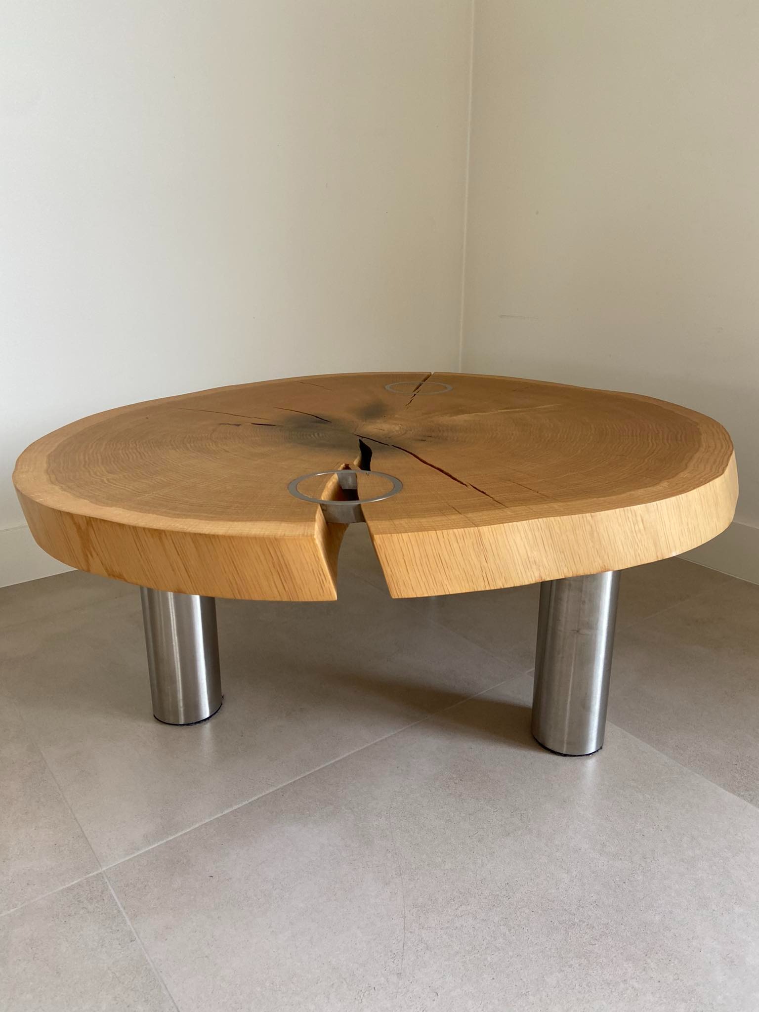 Stolik kawowy z blatem z litego drewna dębowego z charakterystycznymi pęknięciami i metalowymi wstawkami, osadzony na trzech cylindrycznych nogach ze stali nierdzewnej.