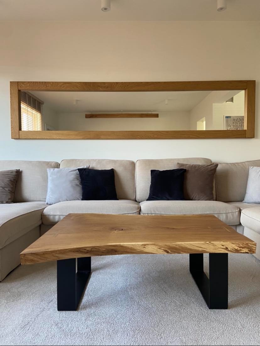 Salon z beżową sofą, drewnianym stolikiem kawowym z czarnymi nogami i dużym, prostokątnym lustrem w drewnianej ramie nad sofą.