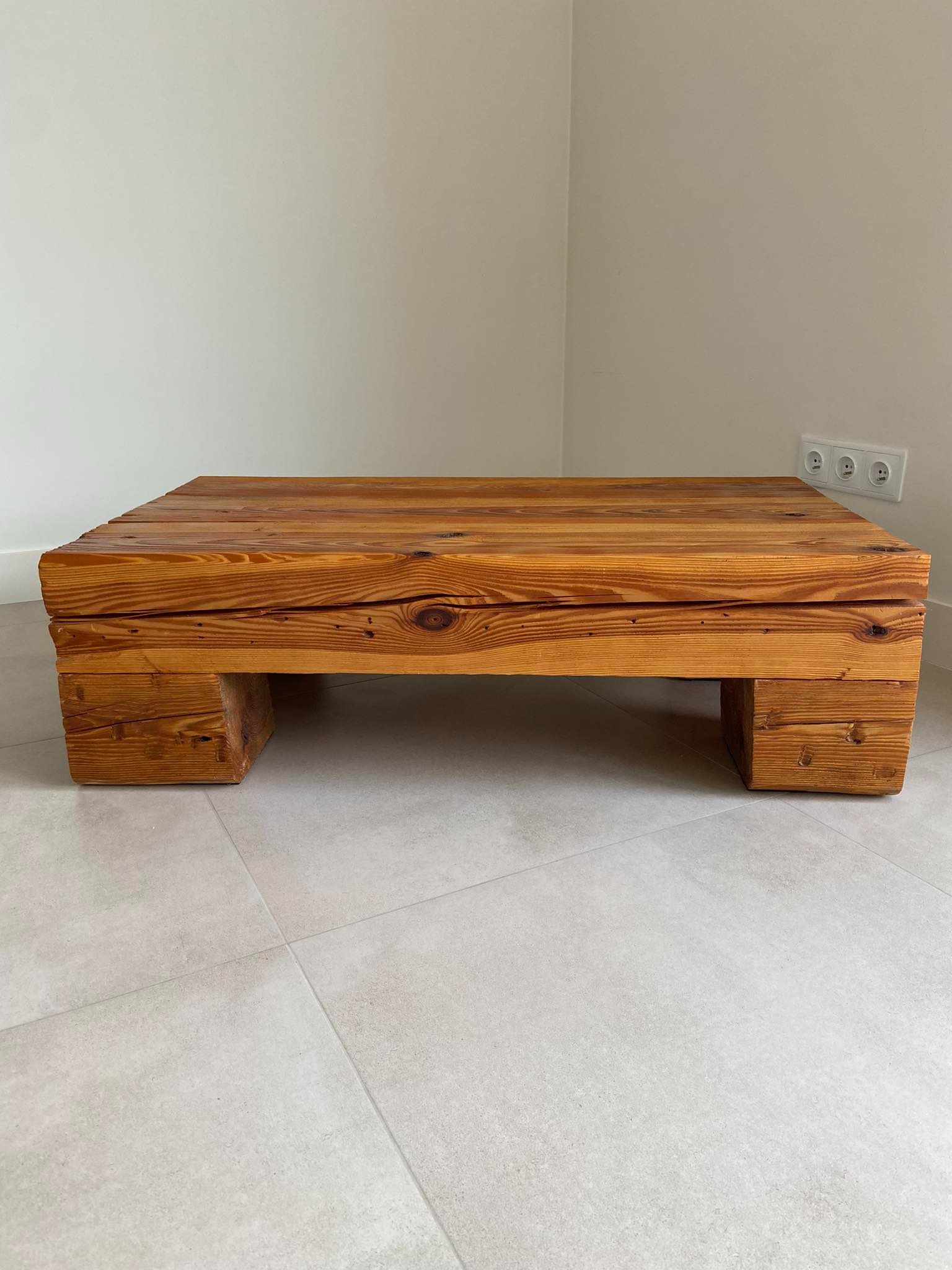 Stolik kawowy z surowego drewna, wykonany z grubych desek i wsparty na niskich, kwadratowych nogach, widoczny na tle szarej podłogi i jasnej ściany z gniazdkiem elektrycznym.