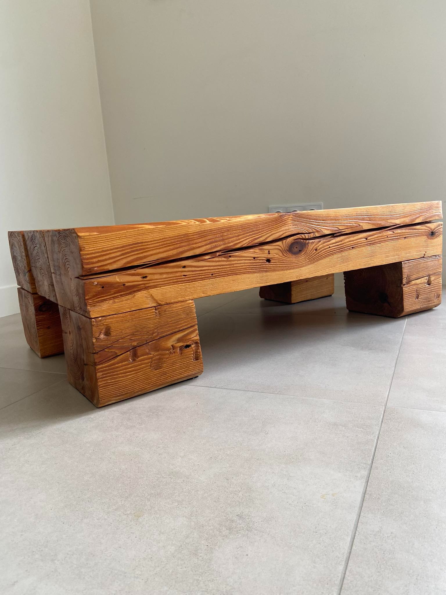 Rustykalny drewniany stolik kawowy z grubo ciosanych belek, wykończony w ciepłym odcieniu brązu, z widocznymi słojami i sękami, ustawiony na szarej podłodze z płytek.