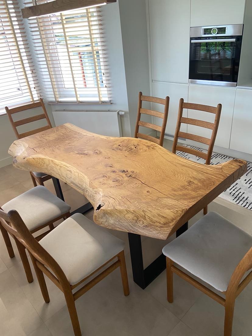 Stół z blatem z naturalnego drewna o nieregularnym kształcie i czarnych, metalowych nogach typu V, otoczony drewnianymi krzesłami z tapicerowanymi siedziskami, na tle jasnej kuchni z zabudową...