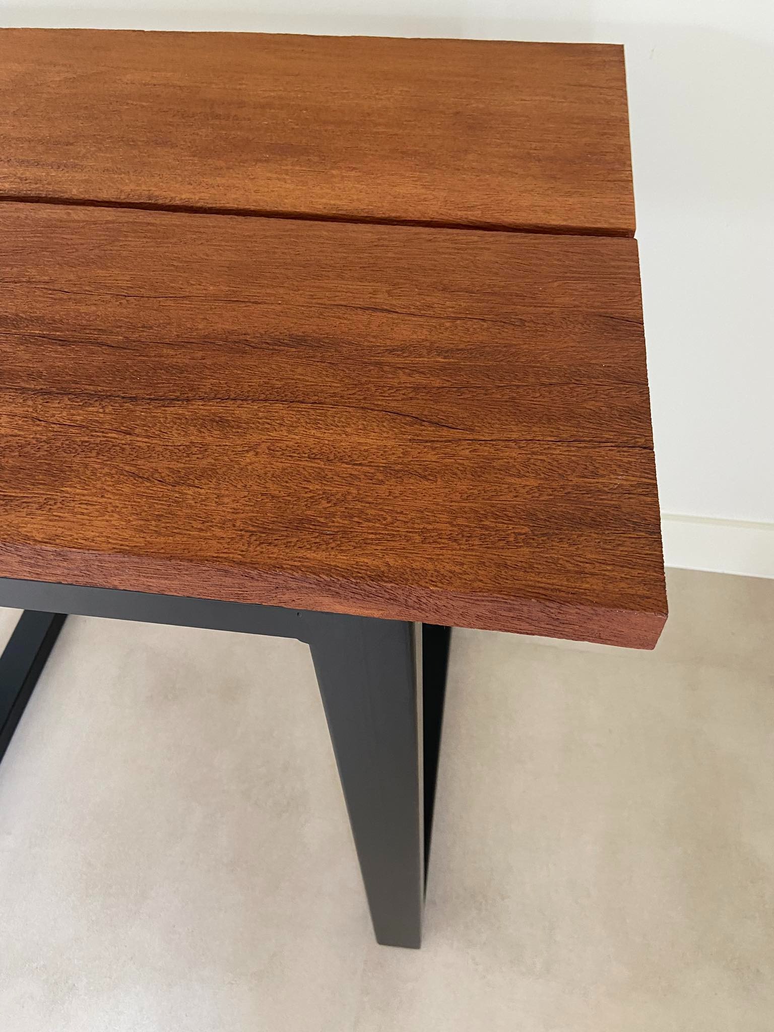 Fragment blatu stołu z egzotycznego drewna o ciepłym, brązowo-czerwonym odcieniu, połączonego z czarnymi, metalowymi nogami w minimalistycznym stylu, na tle jasnej ściany i podłogi.