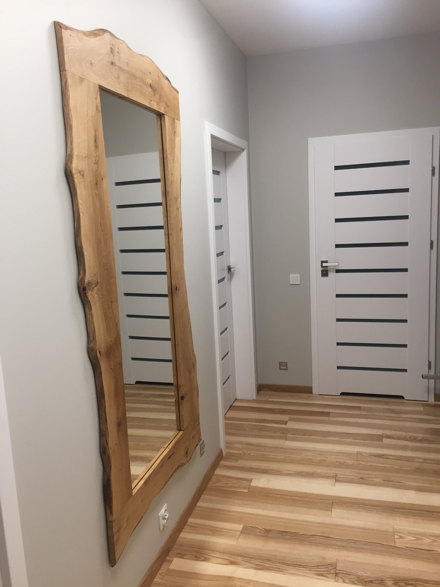 Rustykalne lustro w drewnianej ramie z naturalnymi krawędziami, wiszące w przedpokoju z drewnianą podłogą i białymi drzwiami z poziomymi przeszkleniami.