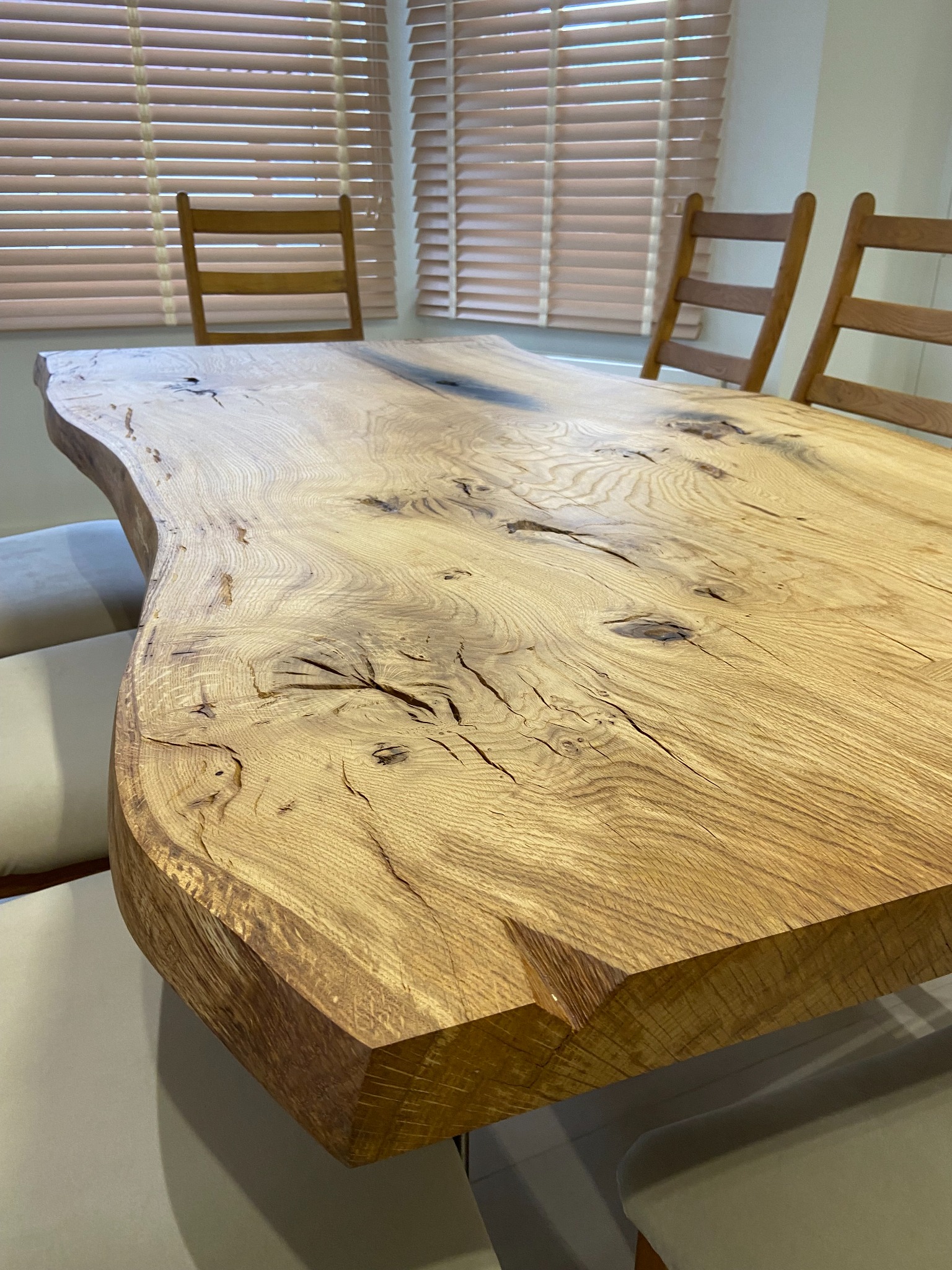 Blat stołu z surowego drewna o nieregularnych krawędziach, z widocznymi sękami i pęknięciami, ustawiony w jasnym wnętrzu z drewnianymi krzesłami i żaluzjami w tle.