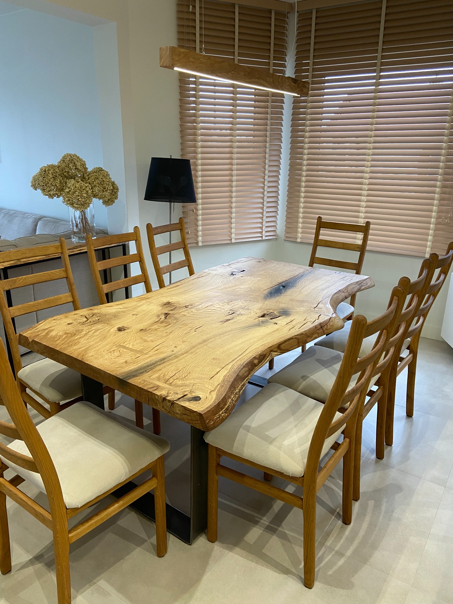 Stół z blatem z litego drewna o nieregularnej krawędzi, wsparty na metalowej podstawie, otoczony drewnianymi krzesłami z jasnymi tapicerowanymi siedziskami, oświetlony drewnianą lampą wiszącą,...