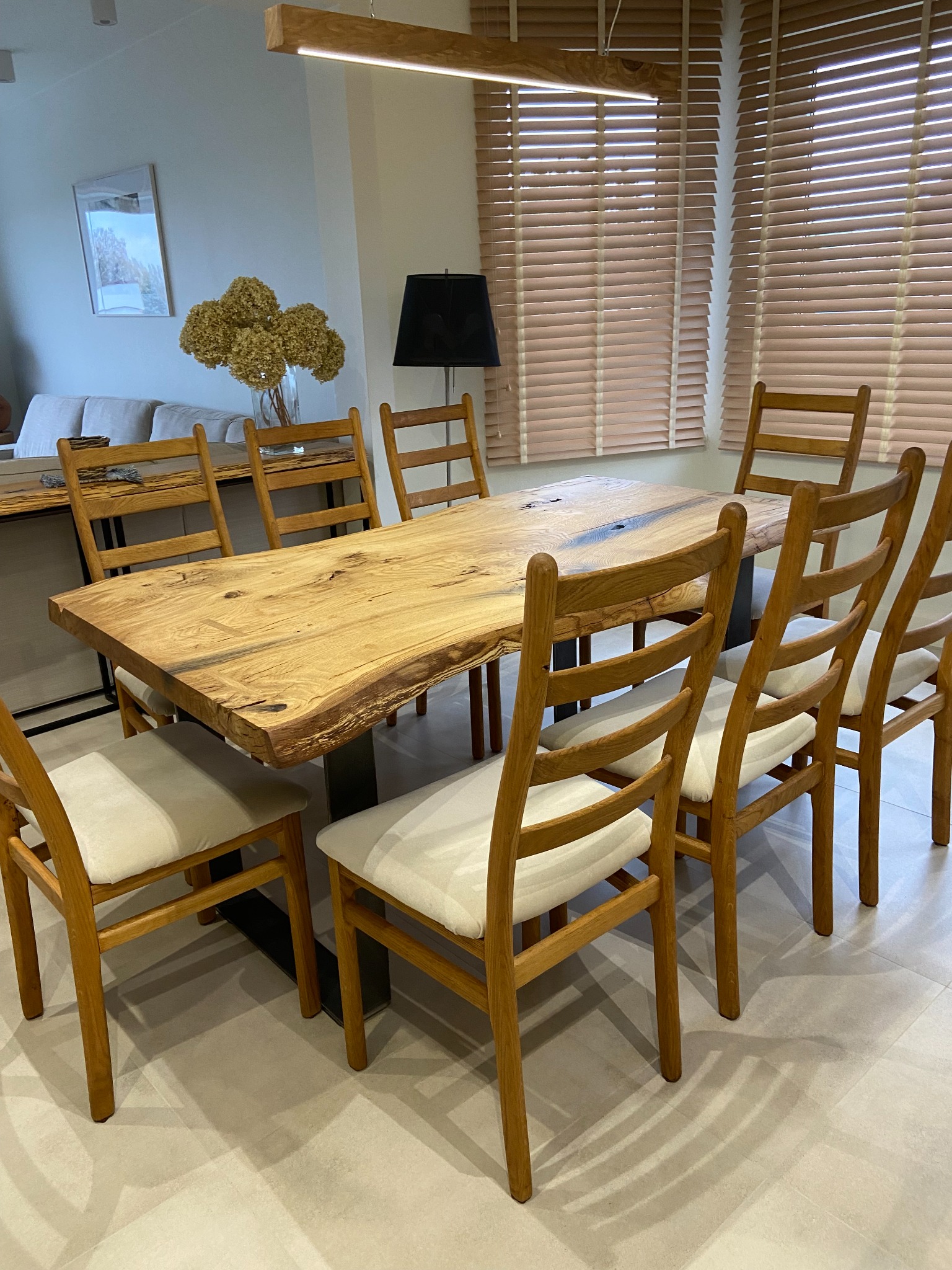 Stół z blatem z litego drewna o nieregularnych krawędziach, wsparty na czarnych, metalowych nogach, otoczony drewnianymi krzesłami z jasnymi siedziskami, w tle żaluzje i dekoracje.