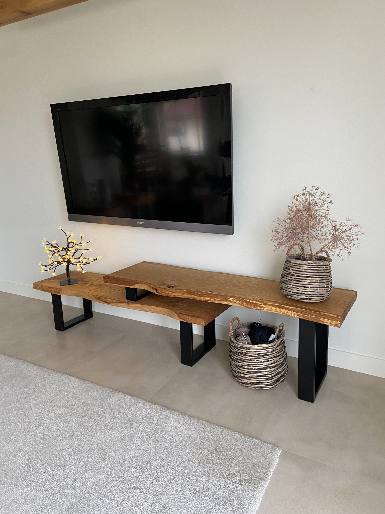 Nowoczesna szafka RTV z drewna z naturalną krawędzią blatu, wsparta na czarnych, metalowych nogach, z dekoracjami w postaci wiklinowych koszy i podświetlanego drzewka, zawieszona pod telewizorem.