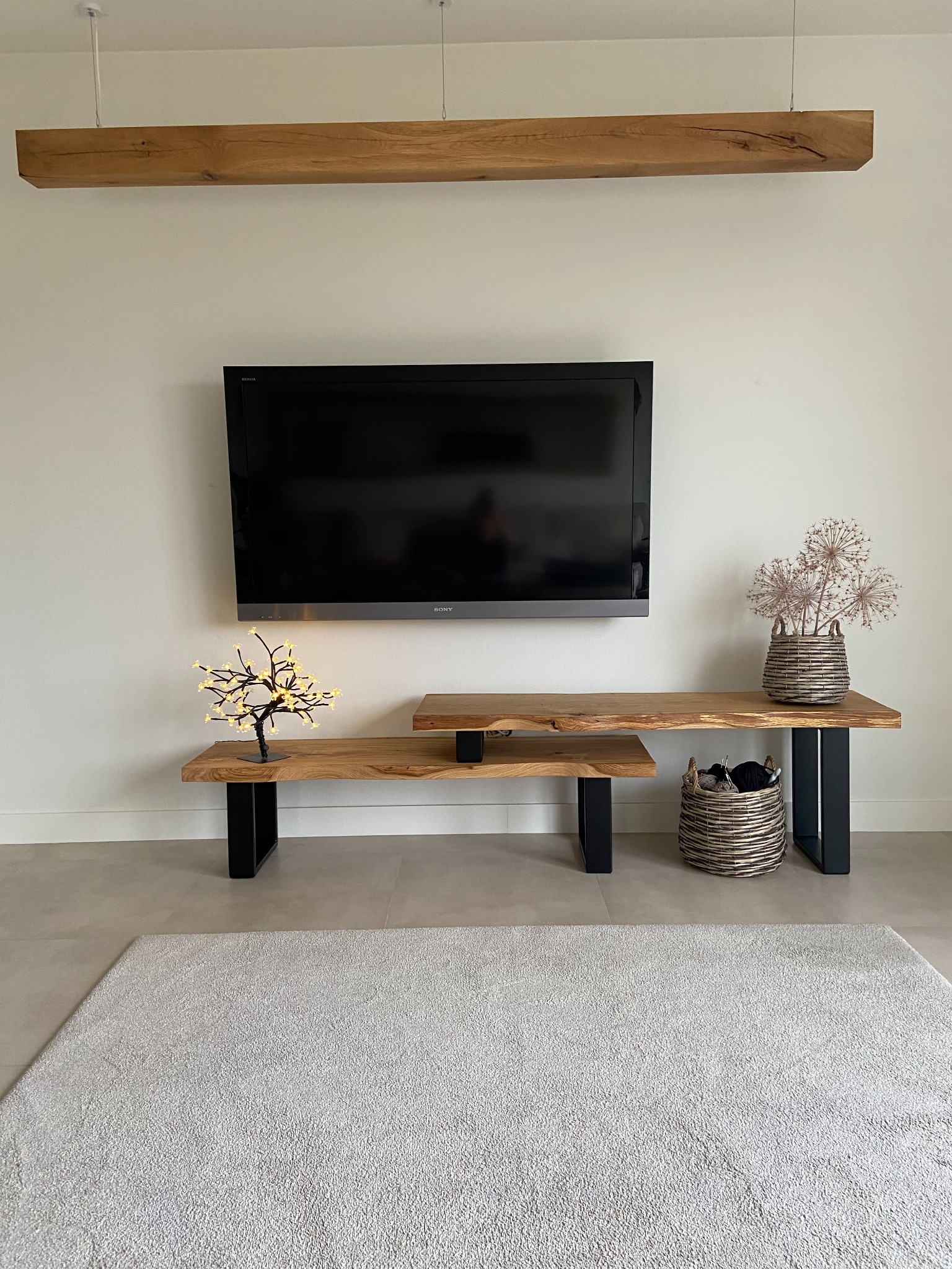 Telewizor zawieszony na ścianie nad drewnianą szafką RTV z blatem o nieregularnej krawędzi, wspartą na czarnych, metalowych nogach. Na blacie dekoracje: podświetlane drzewko i suszone kwiaty...