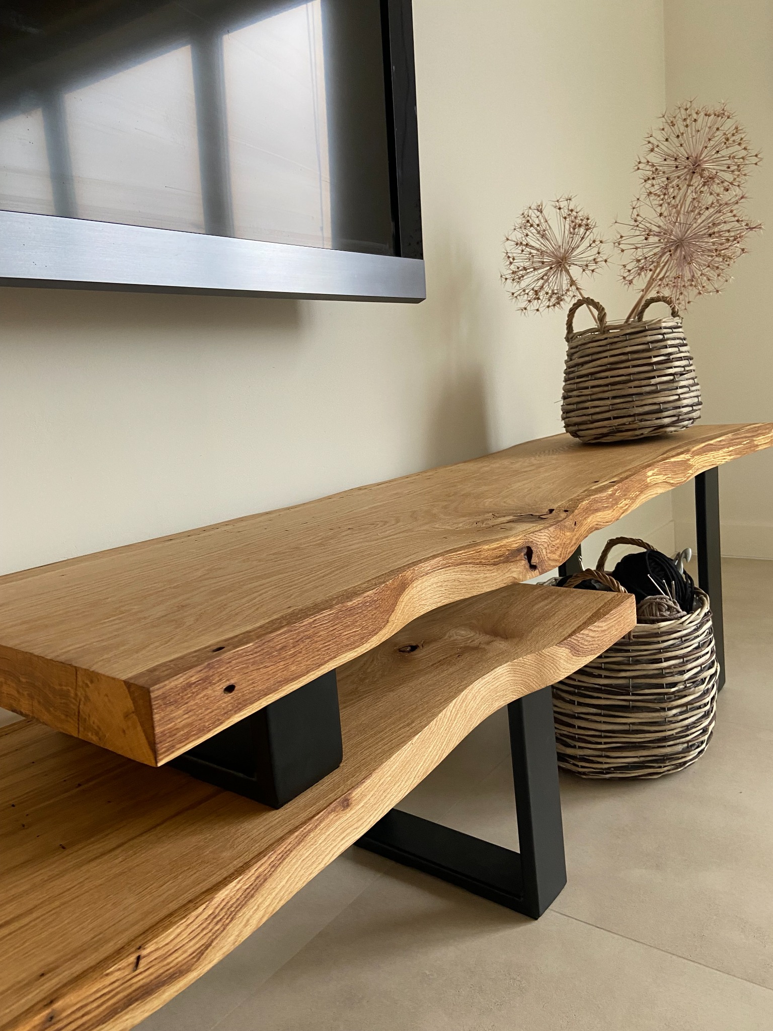 Dwupoziomowa półka pod telewizor z blatem z litego drewna o nieregularnych krawędziach, wsparta na czarnych, metalowych nogach. Na półce wiklinowe kosze z dekoracjami roślinnymi i włóczką.