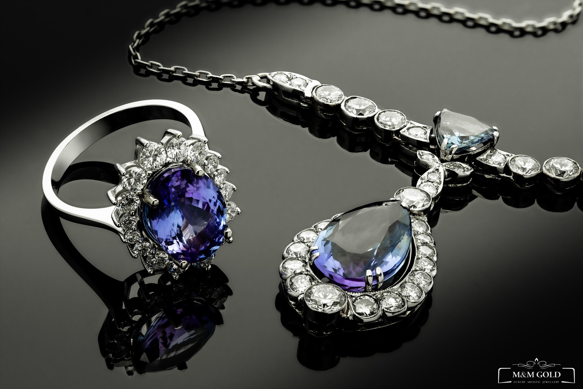 Srebrny pierścionek z dużym, fasetowanym, ametystowym kamieniem otoczonym brylantami oraz srebrny naszyjnik z brylantami i ametystami na czarnym, błyszczącym tle, z widocznym logo firmy jubilerskiej.