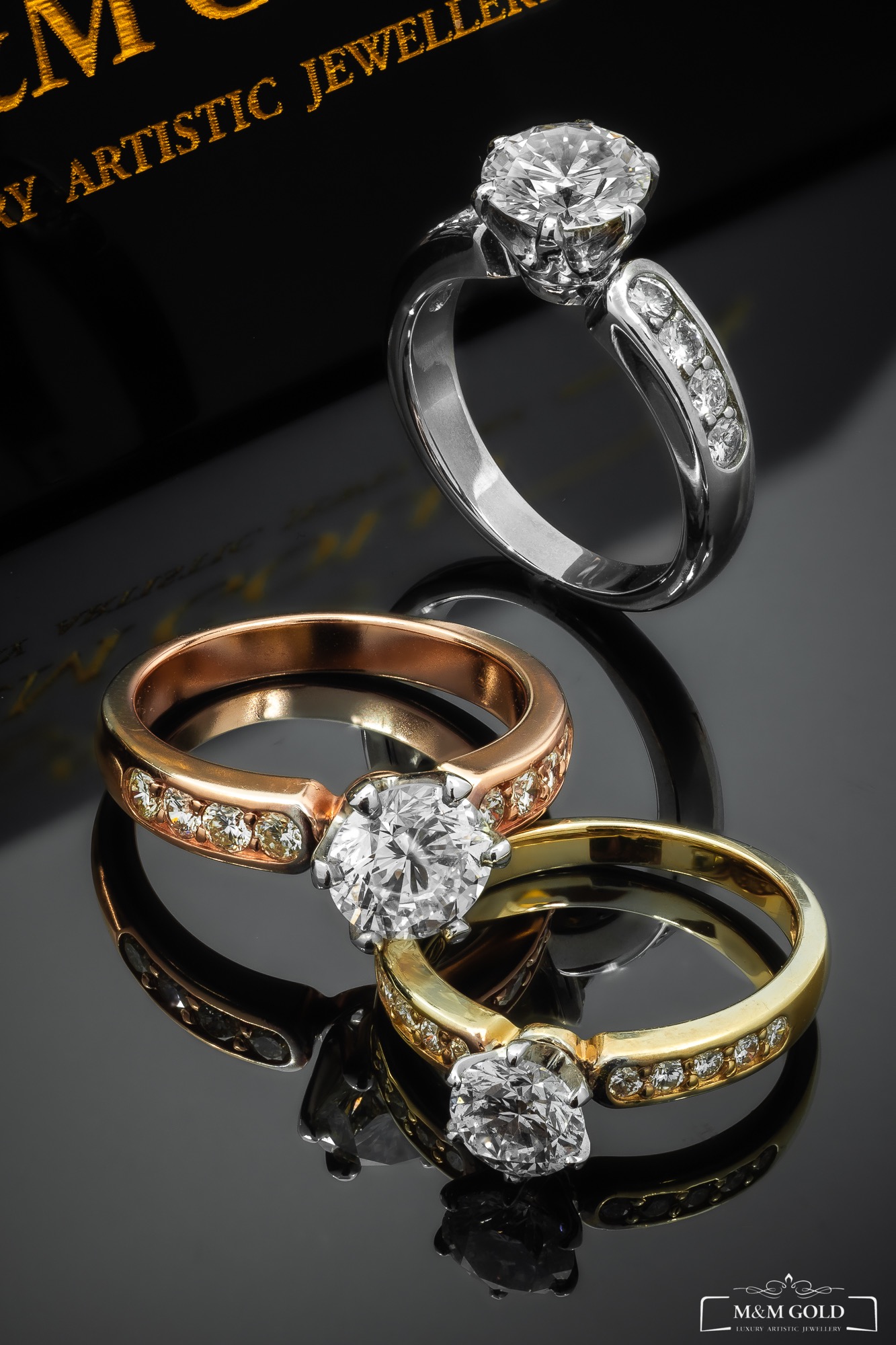 Trzy pierścionki zaręczynowe z dużymi brylantami, wykonane z białego, różowego i żółtego złota, ułożone na czarnej, lustrzanej powierzchni z widocznym logo jubilera.