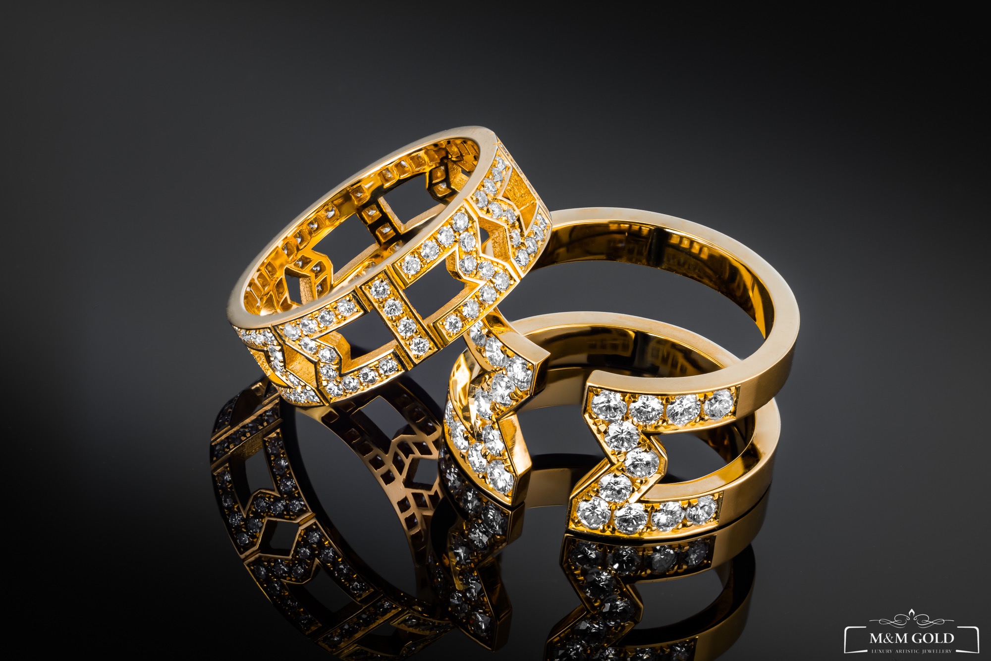 Dwa złote pierścionki z brylantami na czarnym, błyszczącym tle, z widocznym logo M&M Gold w prawym dolnym rogu.