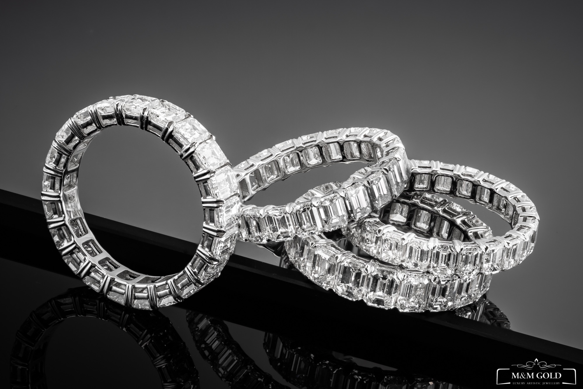 Trzy srebrne obrączki wysadzane prostokątnymi diamentami ułożone na czarnym, błyszczącym tle, tworzące elegancką kompozycję.