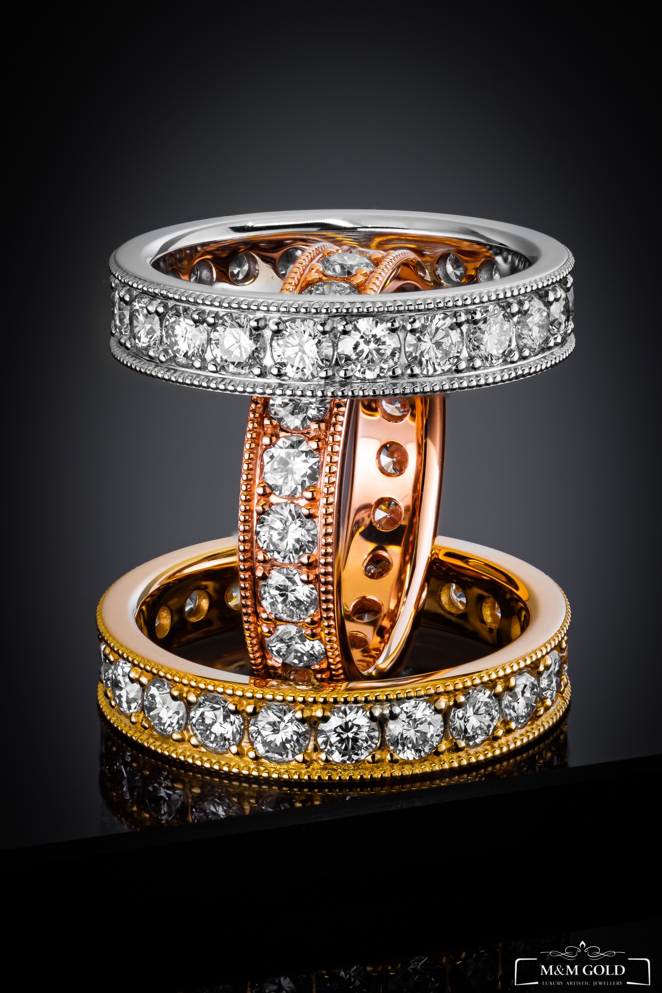 Trzy pierścionki: białe złoto z diamentami na górze, różowe złoto pośrodku z ażurowym wzorem i diamentami, żółte złoto z diamentami na dole, ułożone jeden na drugim na czarnym, błyszczącym tle.