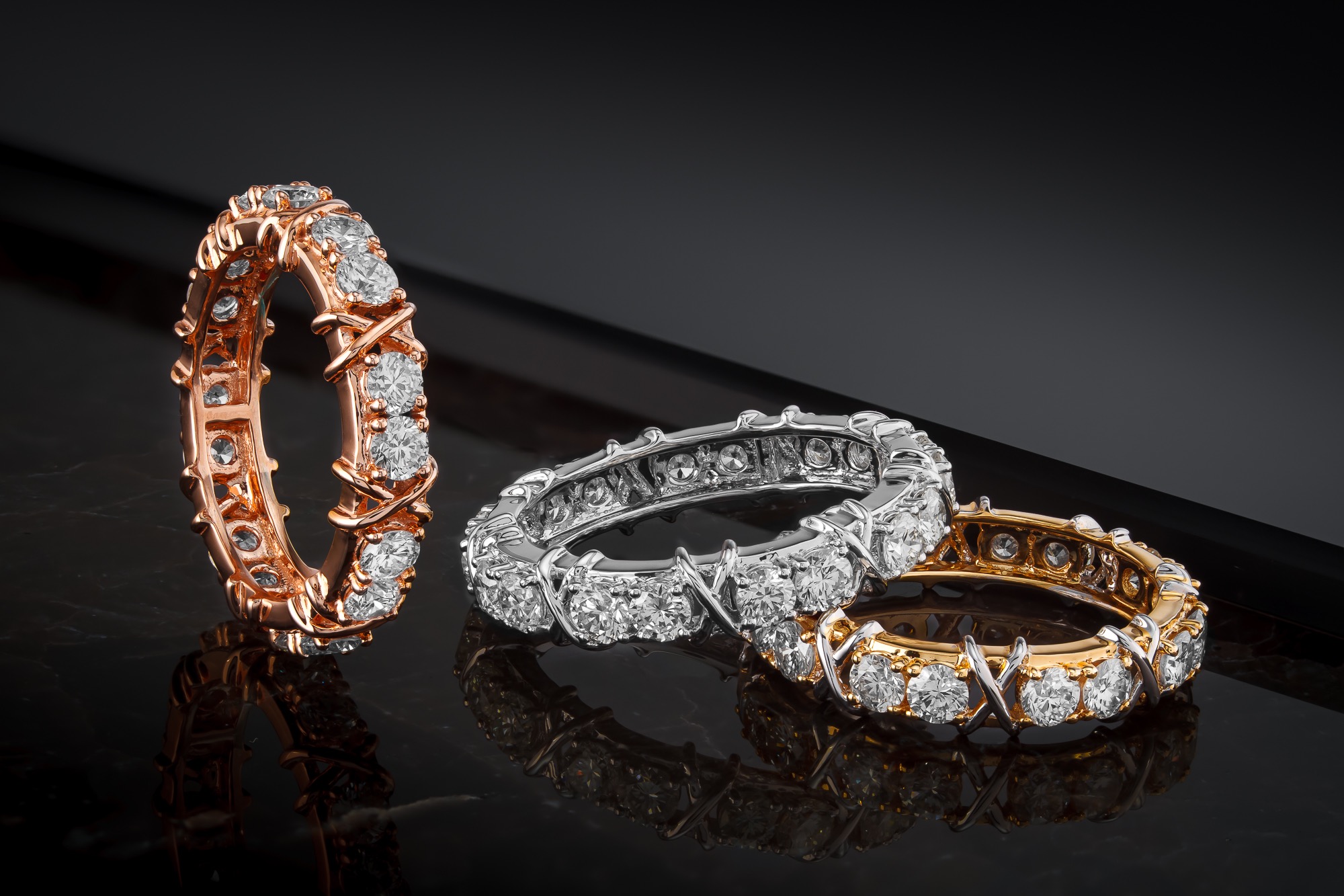 Trzy obrączki wysadzane diamentami: różowe złoto stojące pionowo, białe złoto i żółte złoto leżące na czarnej, błyszczącej powierzchni.