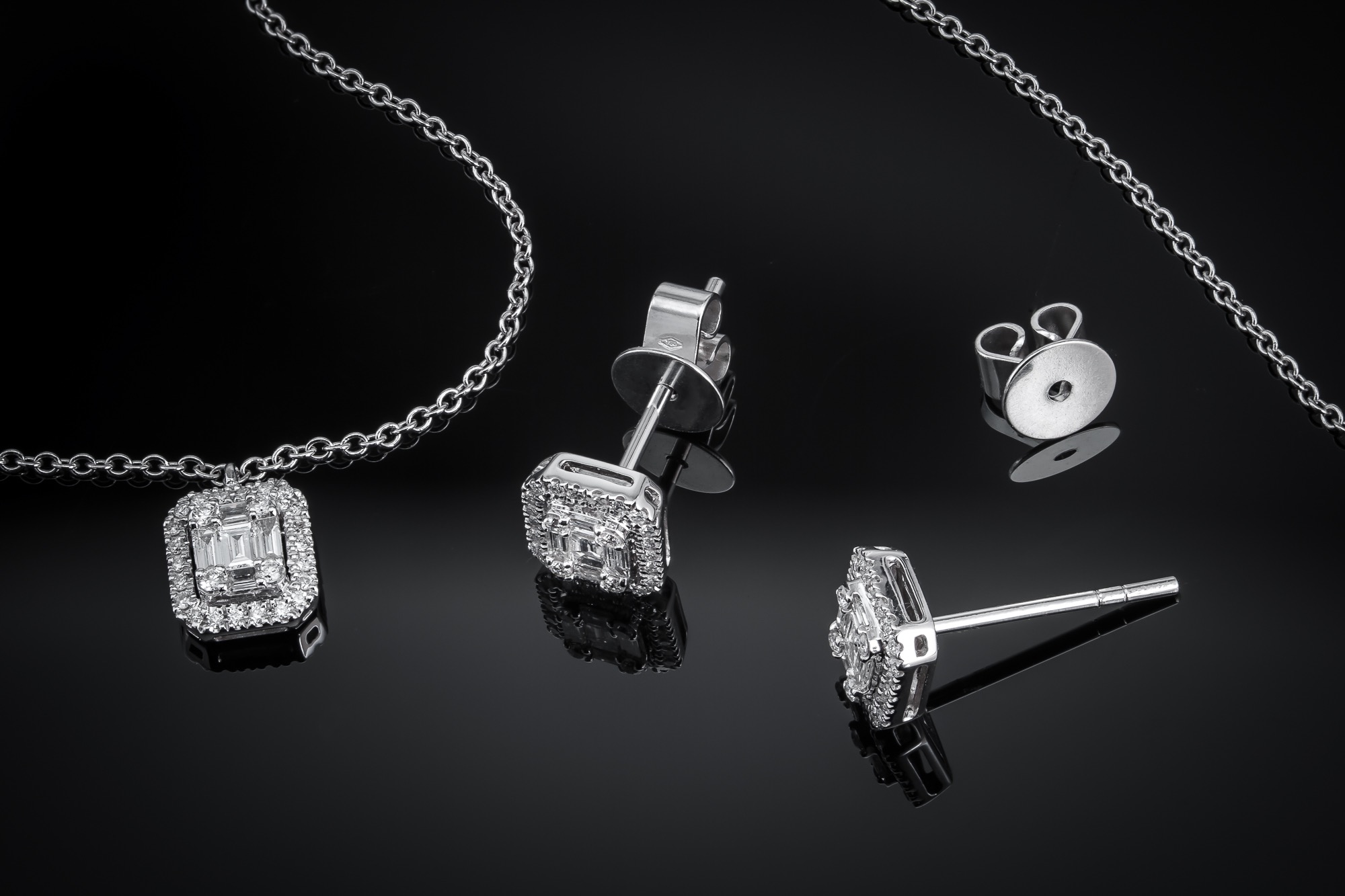 Srebrny komplet biżuterii: kolczyki sztyfty i naszyjnik z zawieszką w kształcie kwadratu z brylantami, na czarnym, błyszczącym tle.
