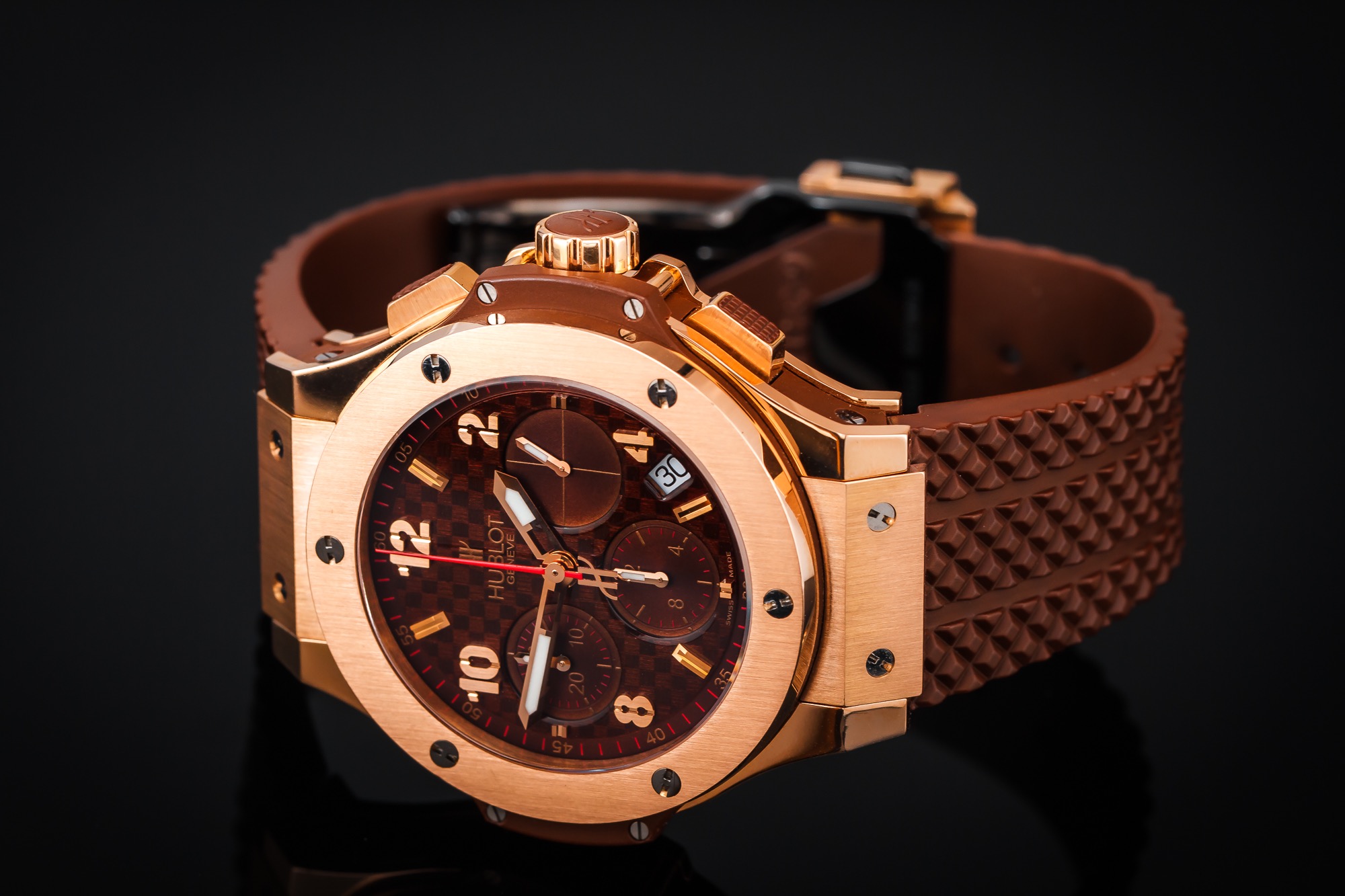 Zegarek Hublot Geneve w kolorze różowego złota z brązową tarczą o wzorze szachownicy i brązowym paskiem z wypustkami, leżący na czarnym, błyszczącym tle.