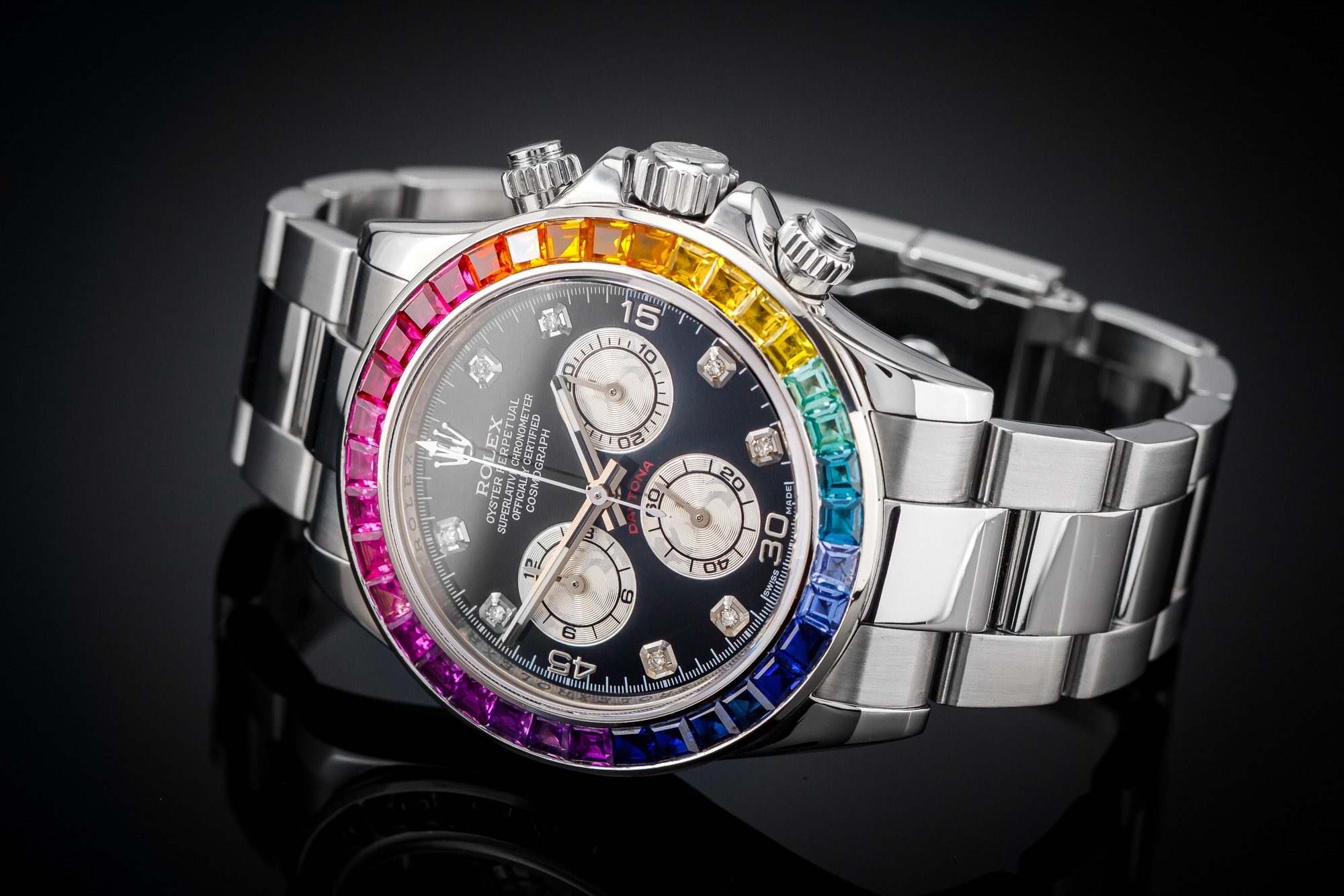 Luksusowy zegarek na bransolecie ze stali nierdzewnej, z czarną tarczą i ramką wysadzaną kolorowymi kamieniami szlachetnymi w układzie tęczy, leżący na czarnym, błyszczącym tle.