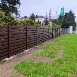 KWK SPAW mobilne naprawy spawalnicze - Długie, poziome, brązowe ogrodzenie panelowe z metalu, oddzielające trawnik od ulicy z chodnikiem, w tle budynki i drzewa.