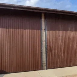 Duża, brązowa brama przesuwna z pionowymi panelami, zamontowana na betonowym fundamencie, z fragmentem zielonej blachy trapezowej po bokach i niebieskim niebem w tle.