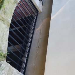 KWK SPAW mobilne naprawy spawalnicze - Zabezpieczone okno piwniczne z metalową kratą, obudowane kamienną ramą, widoczne elementy izolacji i rura odprowadzająca wodę.