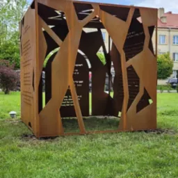 Rdzawa, ażurowa konstrukcja metalowa w kształcie sześcianu z wyciętymi wzorami i tekstem, umieszczona na trawniku w przestrzeni miejskiej.