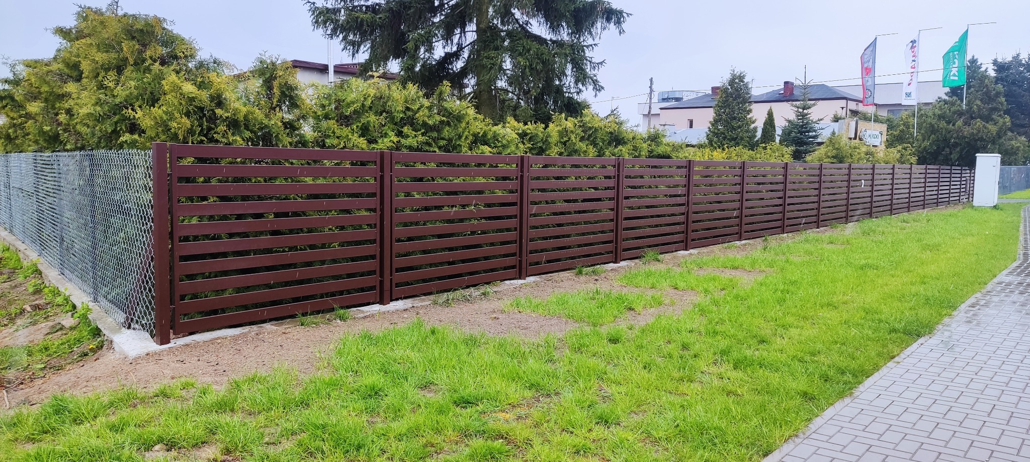 Długie ogrodzenie panelowe w kolorze brązowym z poziomymi szczeblami, przylegające do siatki ogrodzeniowej, otoczone zielenią i kostką brukową.