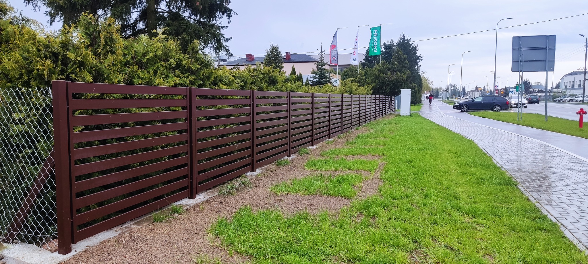 Długie, poziome, brązowe ogrodzenie panelowe z metalu, oddzielające trawnik od ulicy z chodnikiem, w tle budynki i drzewa.