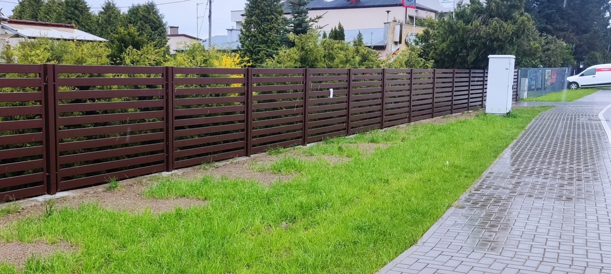 Nowoczesne, poziome ogrodzenie z ciemnobrązowych paneli drewnopodobnych, oddzielające posesję od chodnika z kostki, z widoczną skrzynką elektryczną i fragmentem zaparkowanego samochodu dostawczego...