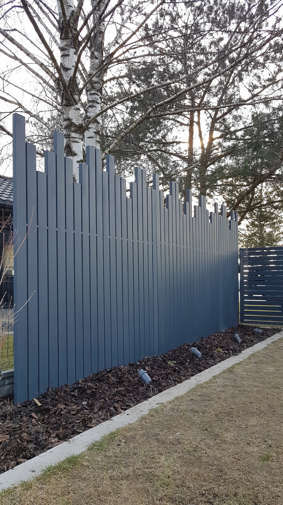 Nowoczesne ogrodzenie panelowe w kolorze grafitowym z pionowych listew o zróżnicowanej wysokości, oświetlone punktowymi lampkami wzdłuż krawędzi rabaty z korą.