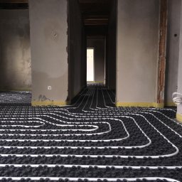 ELEMENTS WARSZAWA - Białe rury systemu grzewczego ułożone na czarnej izolacji w surowym wnętrzu domu jednorodzinnego w trakcie realizacji, widoczne ściany w stanie deweloperskim i żółta folia zabezpieczająca przy...