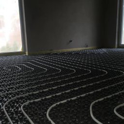 ELEMENTS WARSZAWA - Białe rury ułożone w pętle na czarnej, pokrytej wypustkami powierzchni, tworzące system ogrzewania podłogowego w surowym wnętrzu z oknami.