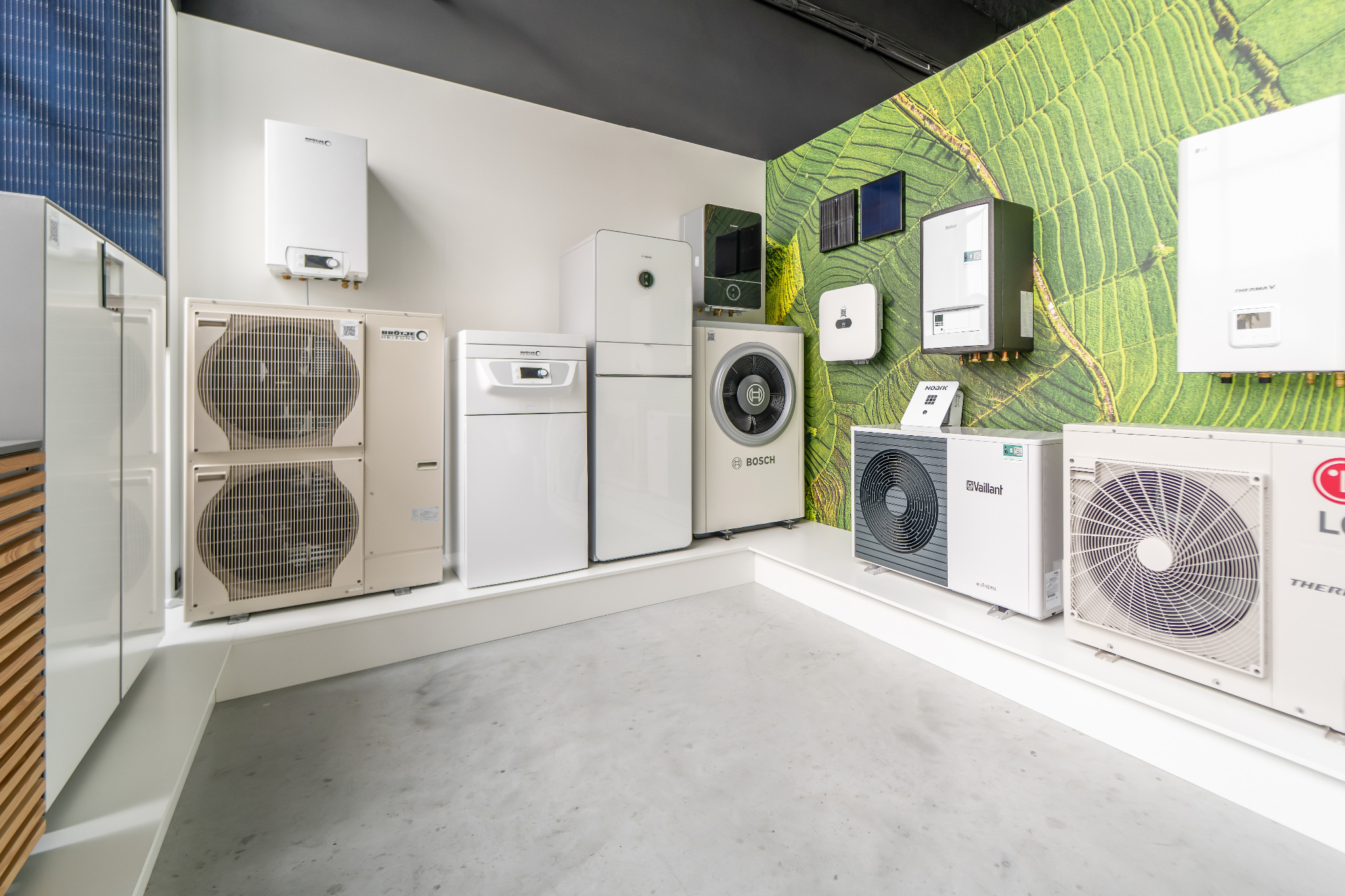 Wystawa różnorodnych urządzeń grzewczych i klimatyzacyjnych w showroomie: pompy ciepła różnych producentów, kotły wiszące i stojące, jednostki zewnętrzne z widocznymi wentylatorami, na tle ściany...