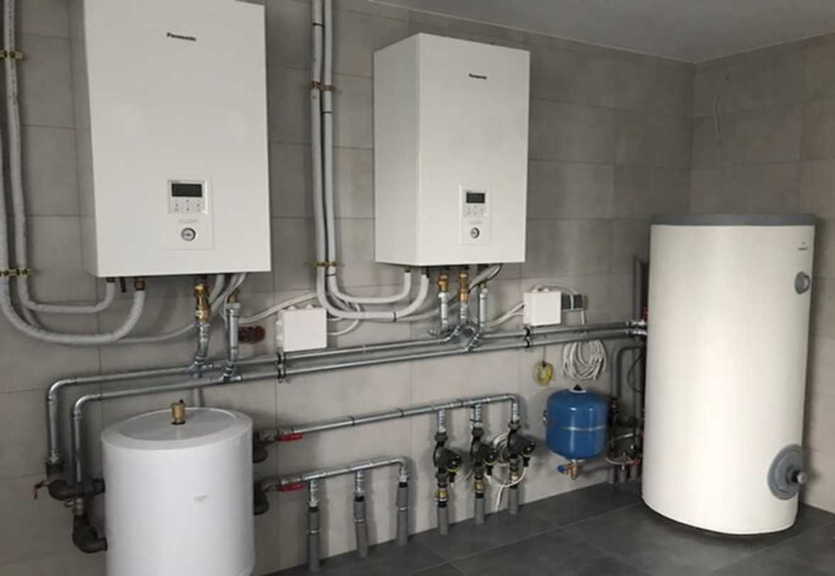 Maszynownia w budynku biurowym z dwoma białymi pompami ciepła Panasonic na ścianie z szarych płytek, zbiornikiem buforowym, naczyniem przeponowym i rozbudowaną instalacją hydrauliczną z rur stalowych.
