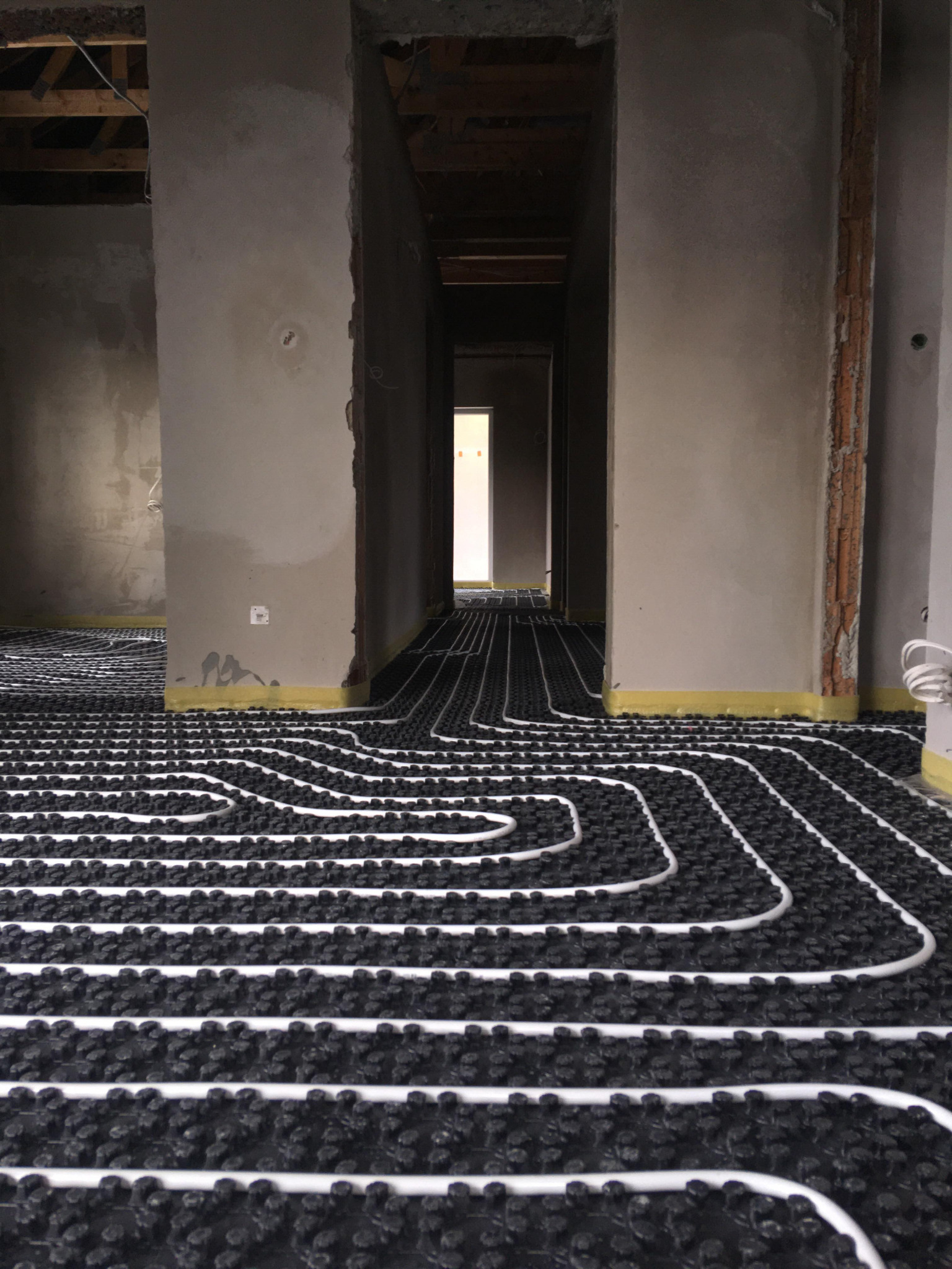 Białe rury systemu grzewczego ułożone na czarnej izolacji w surowym wnętrzu domu jednorodzinnego w trakcie realizacji, widoczne ściany w stanie deweloperskim i żółta folia zabezpieczająca przy...