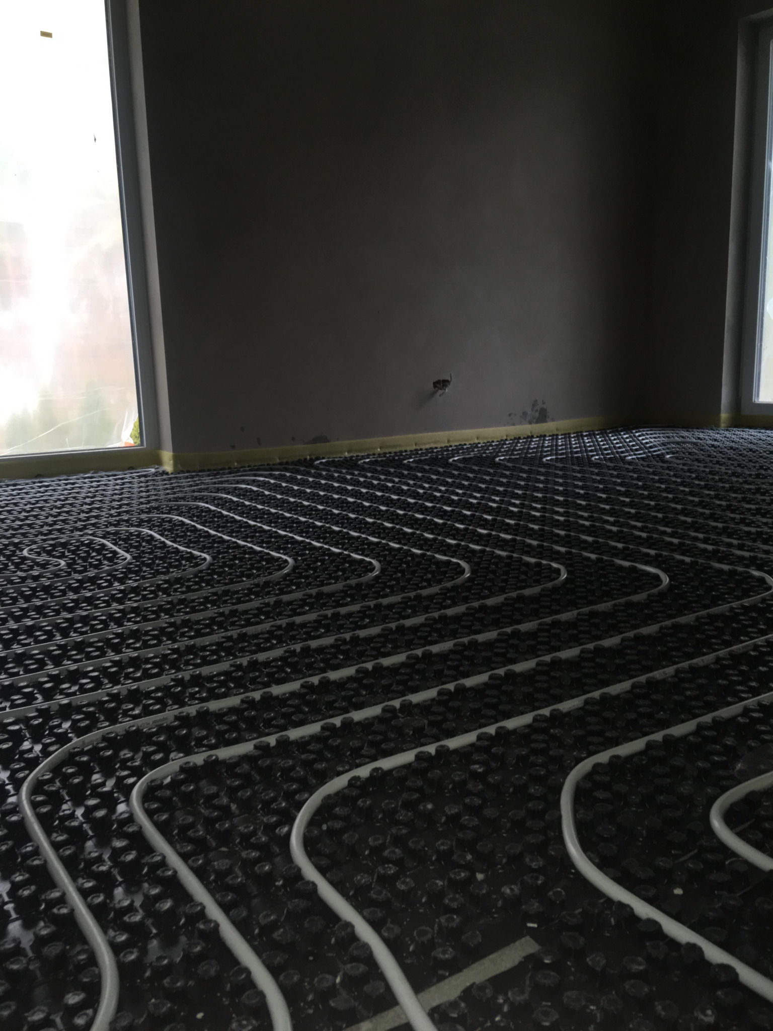 Białe rury ułożone w pętle na czarnej, pokrytej wypustkami powierzchni, tworzące system ogrzewania podłogowego w surowym wnętrzu z oknami.