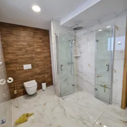 Nowoczesna łazienka z kabinami prysznicowymi ze szklanymi drzwiami, toaletą wiszącą na drewnianej ścianie i marmurową podłogą, w trakcie sprzątania, widoczne ślady prac.
